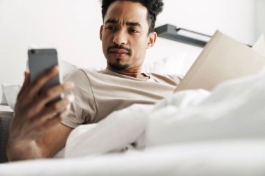 Bıyıklı ciddi bir Afro-Amerikan adamın fotoğrafı. Parlak odada cep telefonu kullanırken kitap okuyor.