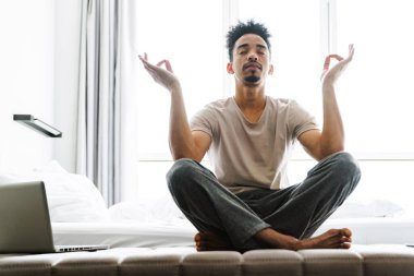 Bıyıklı sakin Afro-Amerikan bir adamın parlak odadaki kanepede otururken meditasyon yaptığı bir fotoğraf.