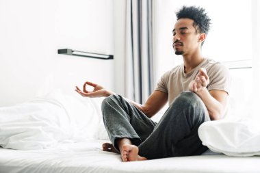 Bıyıklı sakin Afro-Amerikan bir adamın aydınlık odada yatakta meditasyon yaparken çekilmiş fotoğrafı.