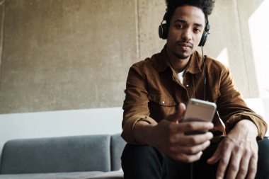 Odaklanmış Afrikalı Amerikalı adamın oturma odasındaki koltukta otururken kulaklık ve cep telefonu kullanmasının fotoğrafı.