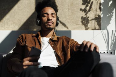 Afro-Amerikan bir adamın oturma odasındaki kanepede otururken kulaklık ve cep telefonu kullandığı fotoğraf.