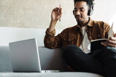 Parlak odada dizüstü bilgisayarla çalışırken kulaklık takan gülümseyen Afrikalı Amerikalı adamın fotoğrafı.