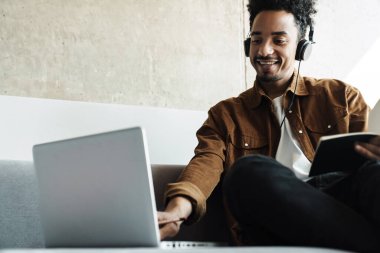 Parlak odada dizüstü bilgisayarla çalışırken kulaklık takan gülümseyen Afrikalı Amerikalı adamın fotoğrafı.