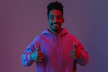 Siyahi kapüşonlu genç Afrikalı Amerikalı adamın portresi. Kamerada zehirleniyor. Başparmakları yukarıda, menekşe arka planında.