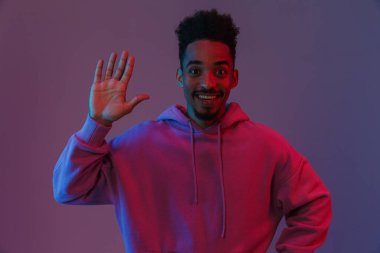 Afro-Amerikan renkli kapüşonlu rahat bir adamın portresi. Kameraya zehirlenmiş, el sallayarak menekşe arkaplanından izole edilmiş.