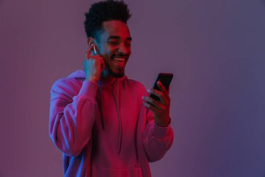 Renkli kapüşonlu Amerikalı neşeli Afro-Amerikalı adamın portresi. Cep telefonuyla konuşuyor. Menekşe arka planında izole edilmiş bir kulaklık var.