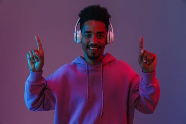 Neşeli bir Afrikalı Amerikalı adam portresi. Kulaklıkla müzik dinliyor ve menekşe arka planında izole bir şekilde parmaklarını yukarı kaldırıyor.