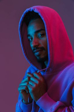 Siyahi kapüşonlu zeki Afro-Amerikan adamın portresi. Mor arka planda izole edilmiş kameraya bakıyor ve poz veriyor.