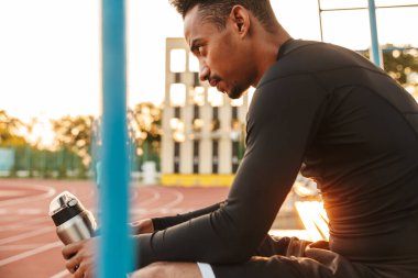 Spor stadyumunda otururken elinde su şişesi tutan Afro-Amerikan bir adamın resmi.