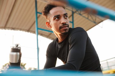 Spor stadyumunda otururken elinde su şişesi tutan kaslı Afro-Amerikan bir adamın görüntüsü