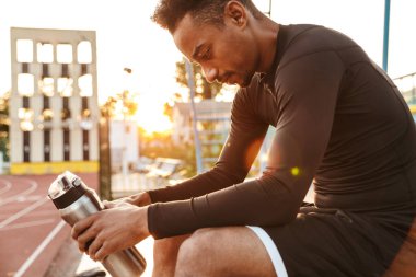 Spor stadyumunda otururken elinde su şişesi olan odaklanmış Afro-Amerikan bir adamın görüntüsü