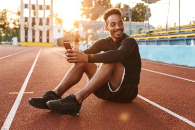 Spor sahasında otururken elinde akıllı telefon tutan Afro-Amerikan bir adamın görüntüsü.