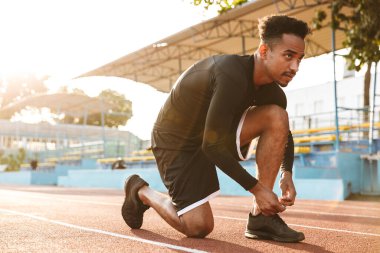 Spor stadyumunda koşarken bağcıklarını bağlayan konsantre Afro-Amerikan bir adamın görüntüsü.