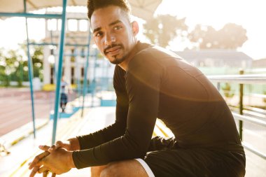 Spor giyimli yakışıklı Afro-Amerikan bir adamın sabah stadyumdaki bankta oturuşu.