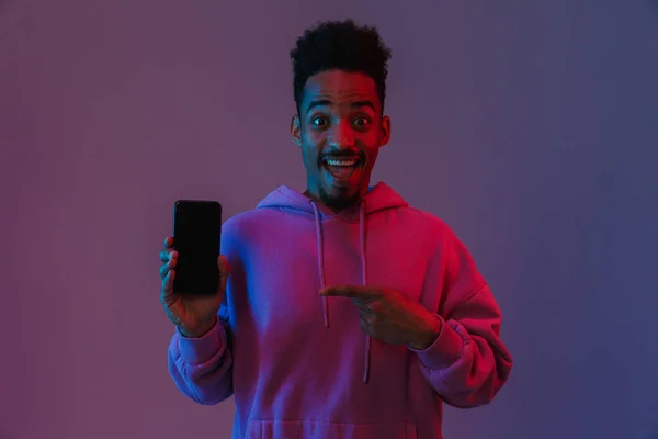 幸せなアフリカ系アメリカ人男性の肖像画でカラフルなパーカーを保持し 指を指すで携帯電話で隔離された紫色の背景 — ストック写真