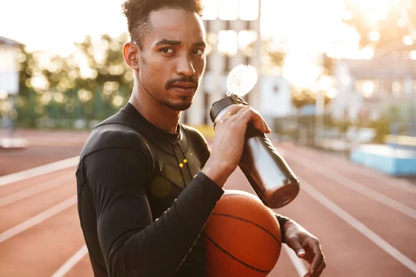 スタジアムの屋外でバスケットボールの飲料水を保持しているアフリカのハンサムな若いスポーツマンのイメージ — ストック写真