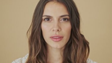 Güzel bir genç kadının yakın plan görüntüsü gözlerini açıyor ve stüdyoda bej bir arka plan üzerinde izole bir şekilde kameraya poz veriyor.