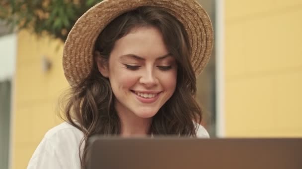 外の居心地の良いカフェでテーブルに座っている間 ラップトップコンピュータを使用して帽子をかぶって幸せな魅力的な女性 — ストック動画