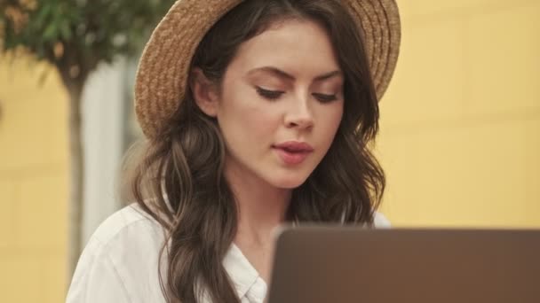 屋外の居心地の良いカフェでテーブルのそばに座っている間 ラップトップコンピュータを使用して帽子をかぶっている無料の魅力的な女性 — ストック動画