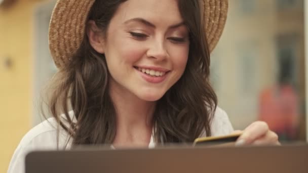 屋外の居心地の良いカフェでテーブルに座っている間 クレジットカードやラップトップコンピュータでオンラインで帽子をショッピングを着て幸せな魅力的な女性 — ストック動画