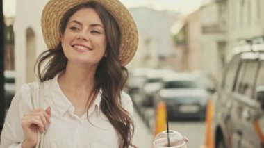 Şapka takan, kahve içen ve sokakta yürürken etrafa bakınan mutlu çekici kadın.