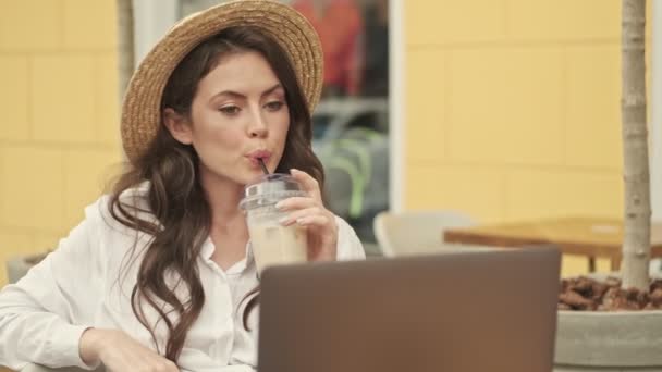 屋外の居心地の良いカフェでテーブルに座っている間 ラップトップコンピュータとコーヒーを使用して帽子をかぶって魅力的な女性を笑顔 — ストック動画