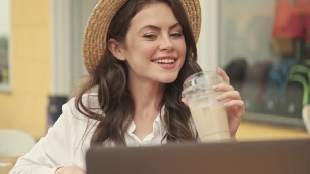 屋外の居心地の良いカフェでテーブルに座っている間 ラップトップコンピュータとコーヒーを飲みながら帽子をかぶって陽気な魅力的な女性 — ストック動画