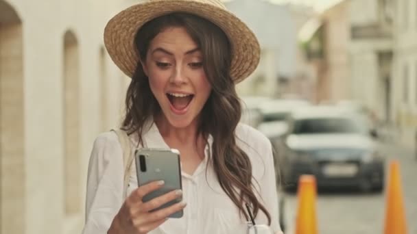 スマートフォンを使って帽子をかぶって路上を歩きながら幸せになる魅力的な女性の笑顔 — ストック動画