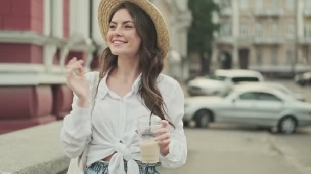 道端でコーヒーを飲みながら歩きながら 帽子をかぶって楽しそうに遊んでいる陽気な魅力的な女性 — ストック動画