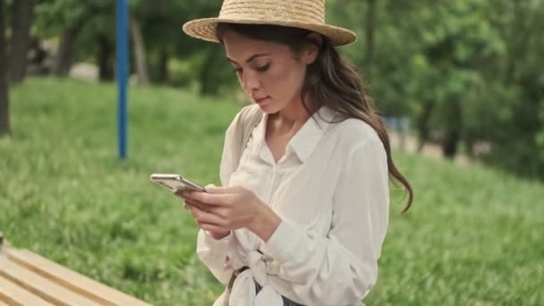 Sorrindo Mulher Atraente Usando Chapéu Usando Smartphone Enquanto Sentado Parque — Vídeo de Stock