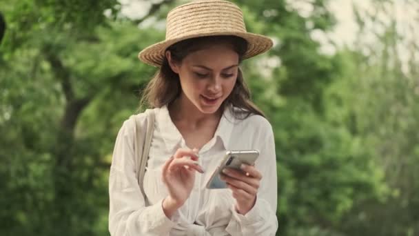 公園に座っている間 スマートフォンを使用して帽子をかぶって満足した魅力的な女性 — ストック動画