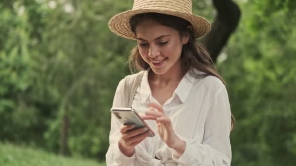 Happy Atrakcyjna Kobieta Nosi Kapelusz Pomocą Smartfona Podczas Siedzenia Parku — Wideo stockowe