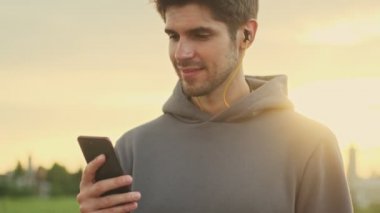 Kulaklıklı, gülümseyen genç bir sporcu dışarıda yürürken akıllı telefonunu kullanıyor.