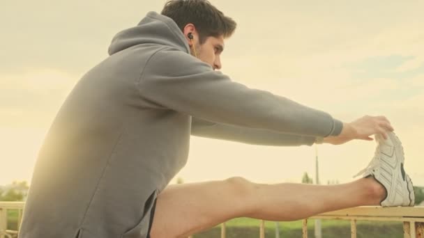 Przystojny Młody Sportowiec Słuchawkami Rozciąga Nogi Zewnątrz Moście — Wideo stockowe