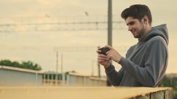 Egy Elégedett Fiatal Sportoló Videót Néz Okostelefonján Miközben Hídon Sétál — Stock videók