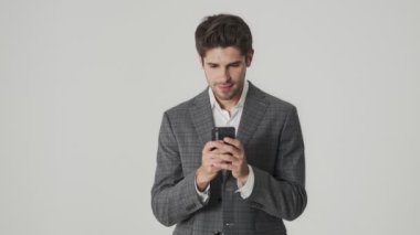 Genç, mutlu, duygusal iş adamı cep telefonu kullanırken beyaz duvar arkaplanından izole edilmiş ve kazanan hareketi gösteriyor.