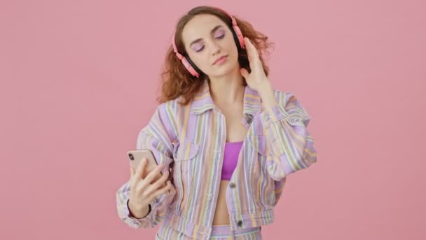 スタジオでピンクの背景に隔離された自撮り写真を撮りながら ヘッドフォンを持つ魅力的な若い女性が音楽を聴いています — ストック動画