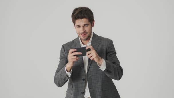 携帯電話でゲームをプレイしながら白い壁の背景に隔離された感情的なビジネスマン — ストック動画