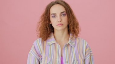 Ciddi bir genç kadın, stüdyoda pembe bir arka planda tek başına duran kameraya baş parmak hareketi yapıyor.