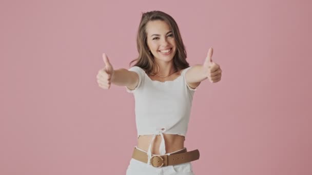 一位快乐的年轻女子站在摄影棚里 双手对着摄像机 在粉色的背景下 摆出一副大拇指的姿势 — 图库视频影像