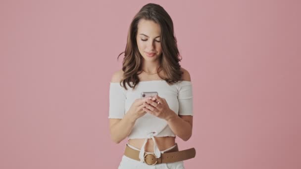 一位心情愉快的年轻女士站在工作室的粉红背景下 在智能手机上打字 — 图库视频影像