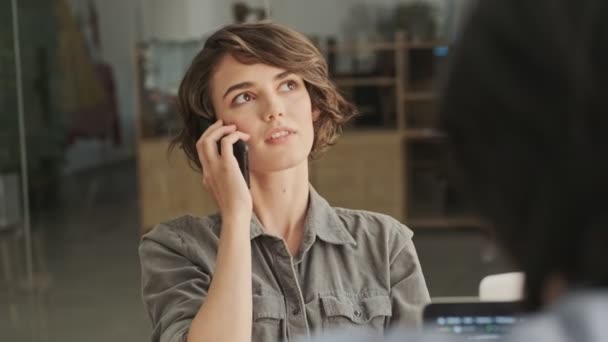 快乐漂亮的女人 穿着衬衫 靠智能手机说话 坐在办公室的桌子旁边环顾四周 — 图库视频影像