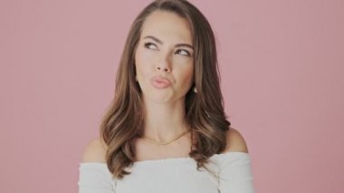 Düşünceli genç bir kadın omuzlarını silkiyor. Stüdyoda pembe arka planda tek başına duruyor.