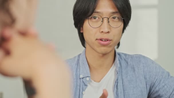 Nyugodt Fiatal Ázsiai Férfi Beszél Valamiről Kollégájával Miközben Asztalnál Irodában — Stock videók