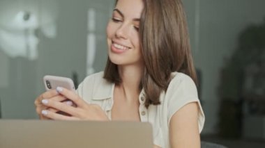 Ofiste dizüstü bilgisayarla otururken akıllı telefon kullanan güzel, esmer bir kadın.