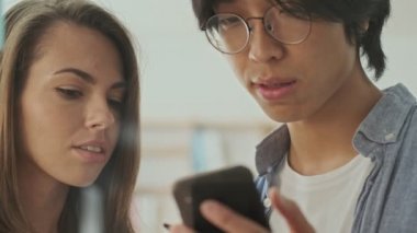 Akıllı telefon kullanan ve ofisteki cam duvara yapıştırılmış renkli çıkartmalara bir şeyler yazan iki sakin genç iş arkadaşı.