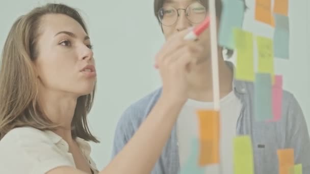 Två Glada Unga Kollegor Skriver Något Färgglada Klistermärken Limmade Glasvägg — Stockvideo