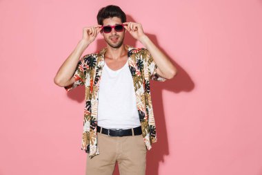 Tıraşsız, güneş gözlüklü yakışıklı bir adamın portresi. Pembe arka planda izole edilmiş kameraya bakıyor.