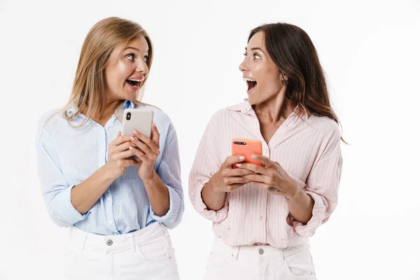 白い背景に隔離された携帯電話を使いながら驚きを表現する興奮した2人の女性のイメージ — ストック写真
