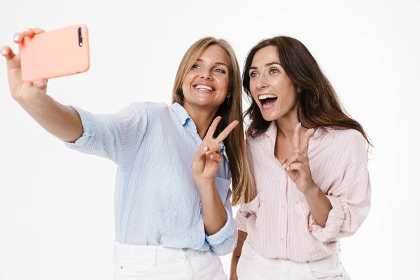 迷人而兴奋的两个女人给和平标志做手势 然后用白色背景的手机自拍的图片 — 图库照片